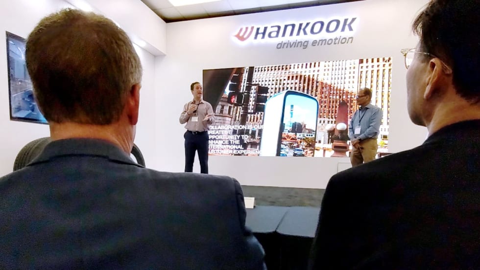 Two presenters speak at a Hankook tire industry event while audience members watch, with a large digital display and illuminated Hankook “driving emotion” logo behind them.