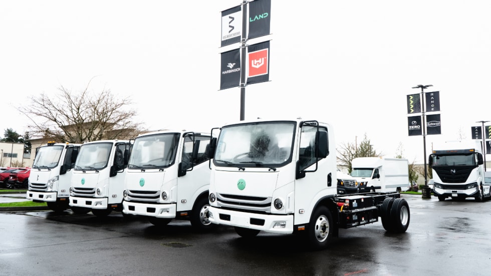 Four white electric cab-over work trucks are parked on wet pavement at a commercial vehicle facility, with additional zero-emission trucks and manufacturer banners visible in the background.