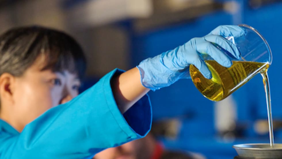 A laboratory technician wearing protective gloves pours a golden liquid from a beaker during testing or analysis in a laboratory setting.