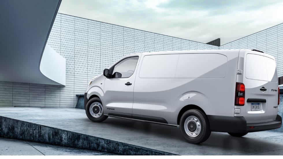 White Ram ProMaster City cargo van shown from the rear three-quarter view outside a modern industrial building, highlighting its windowless cargo area, rear doors, and compact commercial design.