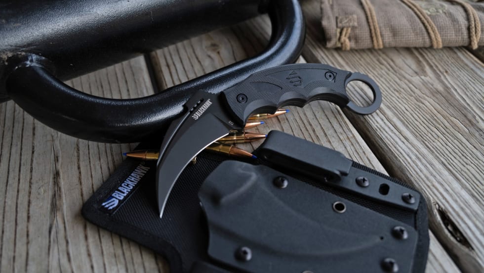 Blackhawk karambit-style fixed-blade knife with finger-ring handle displayed alongside ammunition, a black sheath, and tactical equipment on a wooden surface.