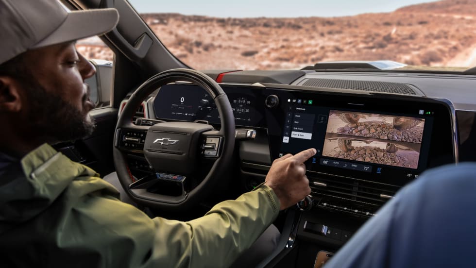Driver inside a Chevrolet Silverado using the center touchscreen to view underbody camera images while parked on rocky terrain.