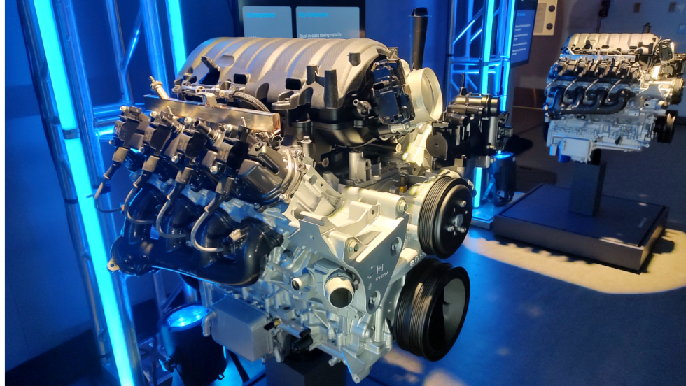 GM small-block V-8 engine displayed under blue lighting, showing intake, ignition, accessory-drive, and exhaust components, with another engine exhibit visible in the background.