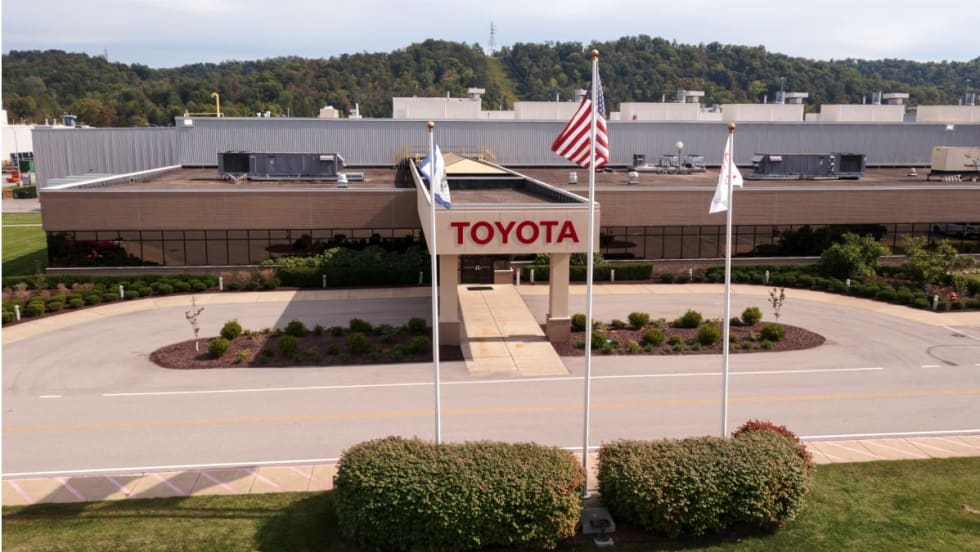 Aerial view of a Toyota manufacturing facility entrance, featuring the Toyota name above the doorway, three flagpoles, landscaped grounds, and the factory complex in the background.
