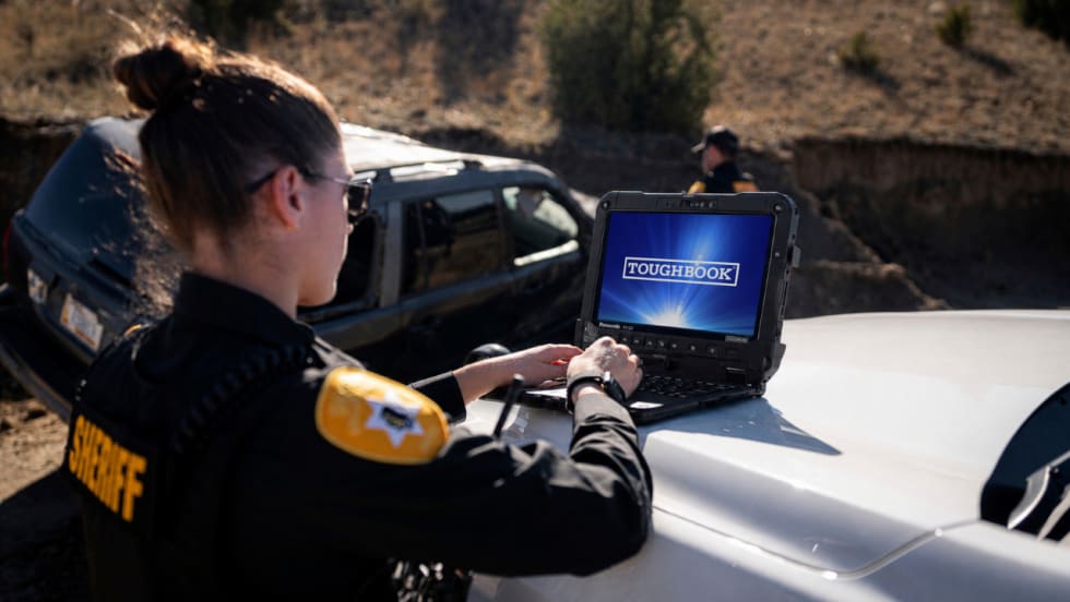 Sheriff's deputy operates a Panasonic Toughbook rugged laptop on the hood of a patrol vehicle at an outdoor law enforcement scene, with another officer and SUV in the background.