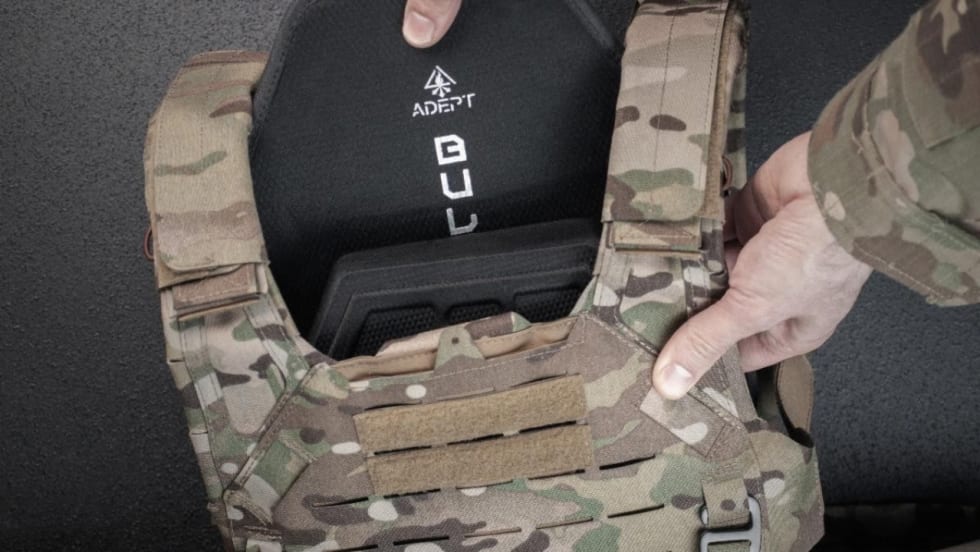Hands insert an Adept Armor protective plate into a camouflage tactical plate carrier, demonstrating how the armor plate fits inside the vest.