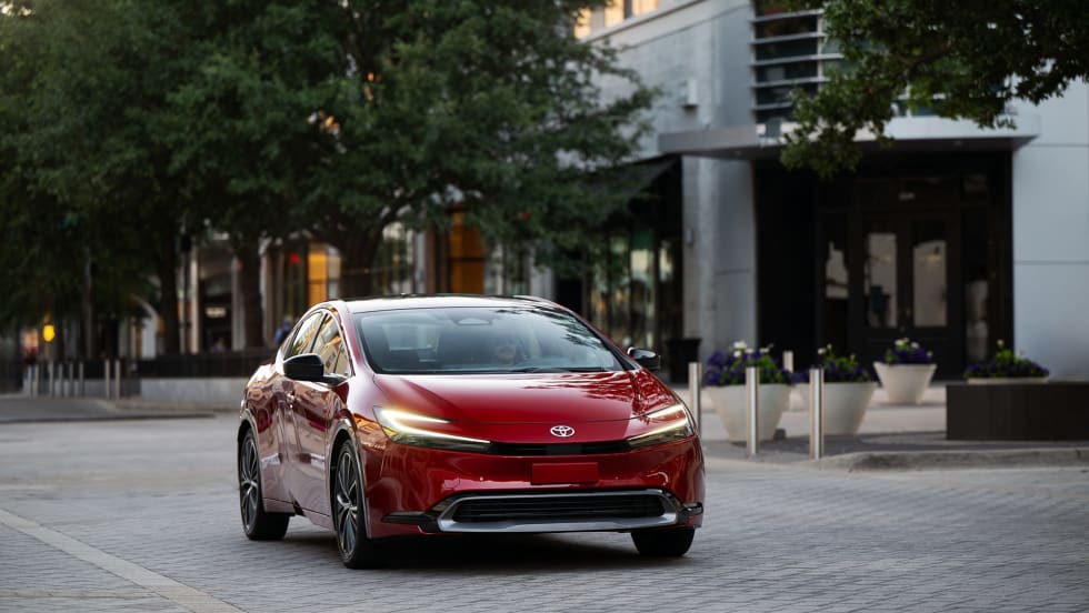 Front three-quarter view of a red 2027 Toyota Prius parked in an urban setting, showcasing the updated hybrid sedan's sleek design, LED lighting and aerodynamic styling.
