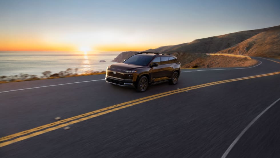 Subaru electric SUV driving along a coastal highway at sunset, representing the 2027 Getaway with three-row seating and all-electric performance.