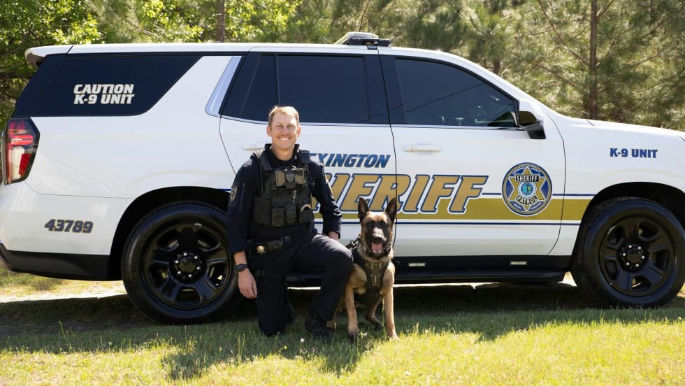 K-9 police officer with German Shepherd beside sheriff's white patrol vehicle