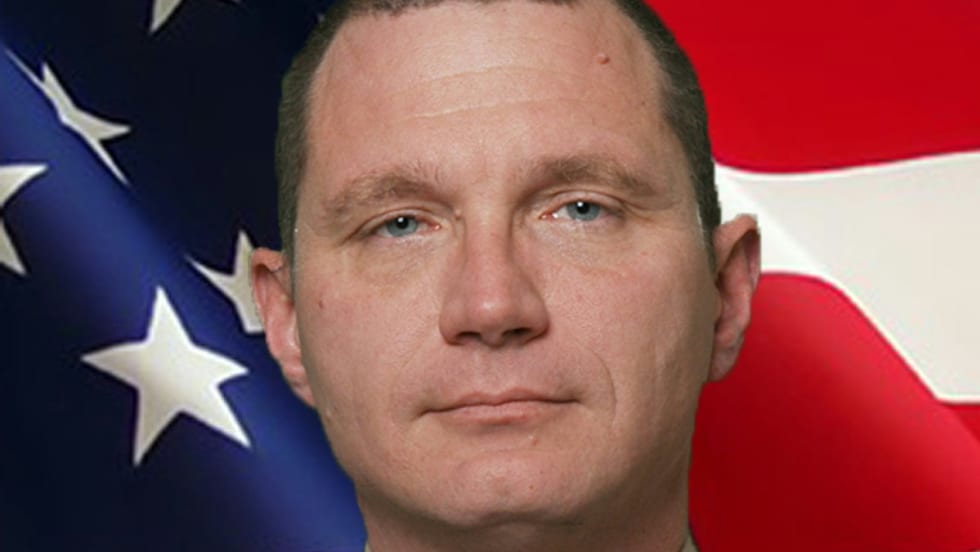 Law enforcement officer in tan uniform with sheriff's badge, standing before American flag