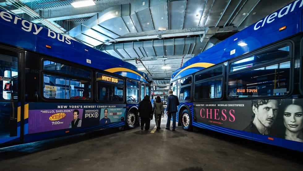 A wide angle view of two MTA buses with three people walking between them.