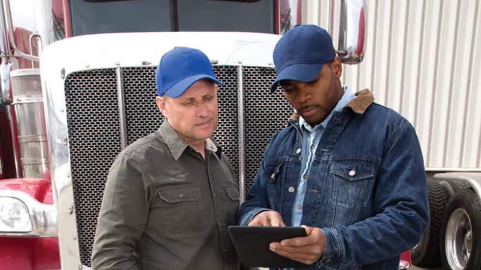 Two truck fleet managers standing in front of a semi-truck reviewing emergency response plans on a digital tablet.