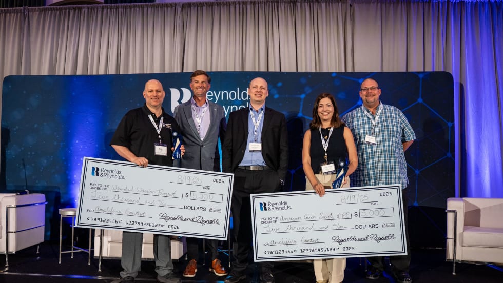 Group of five people on stage holding large checks at a Reynolds and Reynolds event, recognizing winners of an automotive innovation contest.