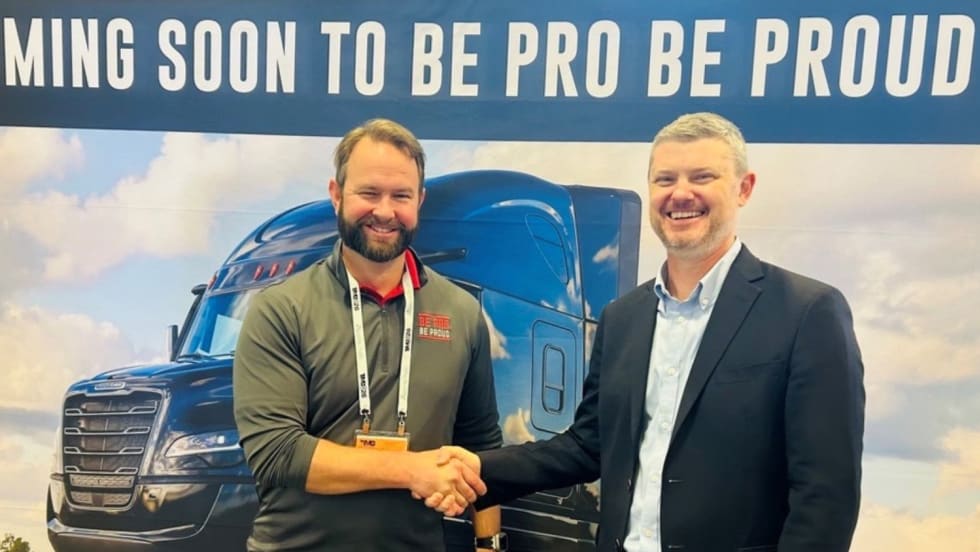 Two men shake hands in front of a Freightliner truck display during a partnership announcement supporting workforce development and training programs.