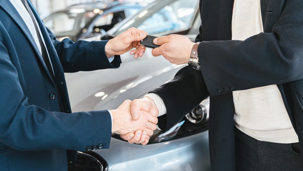 two men shaking hands with one handing over a set of car keys, a car in the background