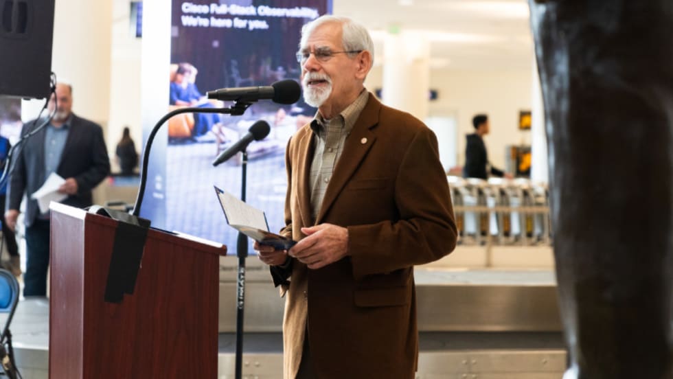 Rod Diridon Sr. speaking at a podium wearing a brown blazer.