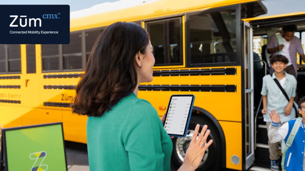 A woman holds a tablet and waves at children disembarking a school bus.