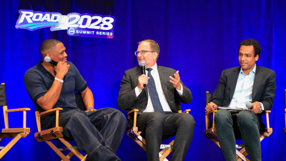 Three speakers participate in a panel discussion at the Road to 2028 Summit Series. A man in a gray suit speaks into a microphone while two other panelists listen on stage beneath a "Road to 2028 Summit Series" sign.