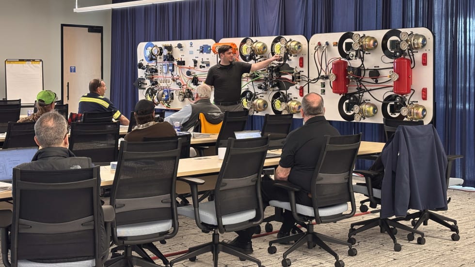 Instructor leads a brake system training class with technicians seated at tables, using a large demonstration board with air brake components and wiring.