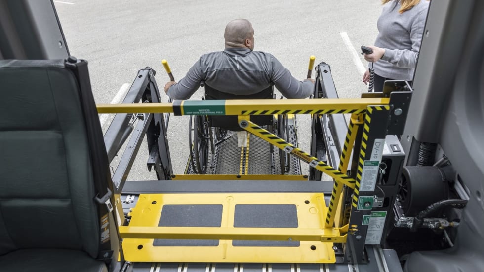 A wheelchair user operates a school bus wheelchair lift equipped with BraunAbility’s LiftSentinel barrier system as another person stands nearby with a handheld control.