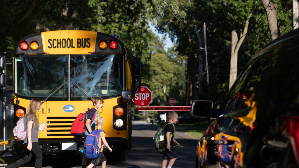 Children cross in front of a stopped school bus with its stop arm extended while a nearby vehicle waits, illustrating school zone safety and risks of illegal passing.