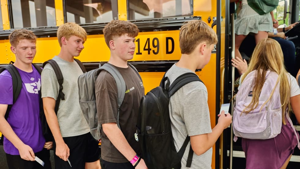 Students wearing backpacks line up to board a yellow school bus, each holding an NFC-enabled student ID card to scan as they enter. The image illustrates student ridership tracking technology that records boarding activity in real time.