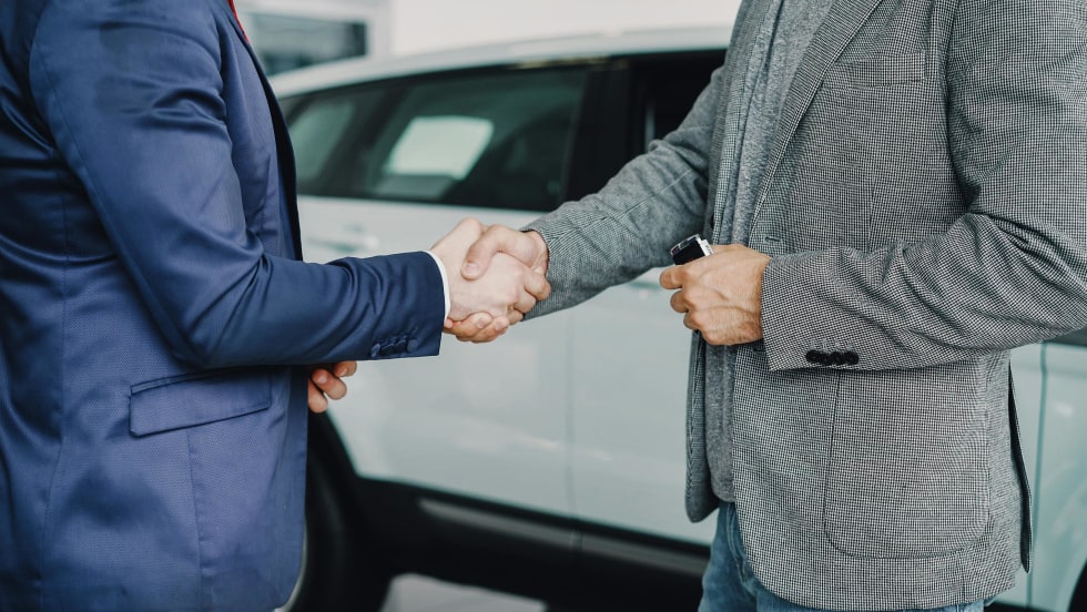 Photo of two men in suit jackets shaking hands next to new car inside of a dealership