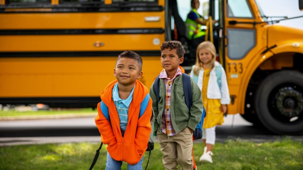 Students walking away from a school bus with a driver in the background, representing efforts to improve student behavior support and safety in school transportation programs.