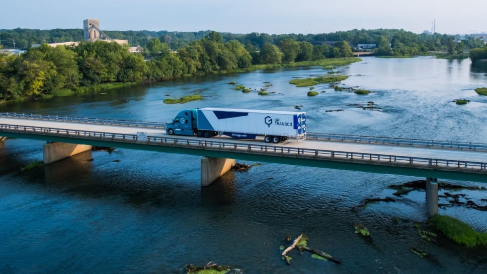 A GP Transco tractor-trailer crosses a bridge over a river, with trailer side skirts visible beneath the white trailer.