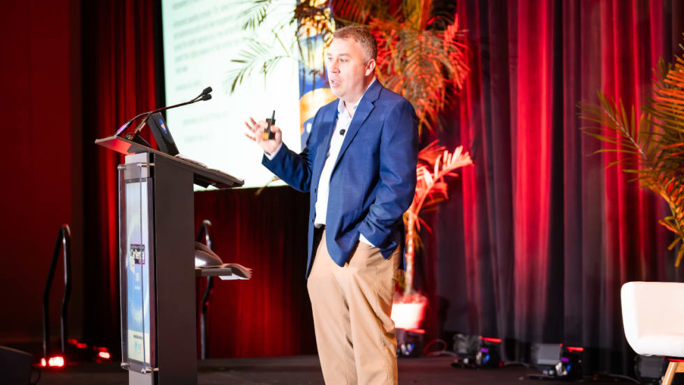 Speaker John Healy on stage with a podium, screen, and red curtains in the background.
