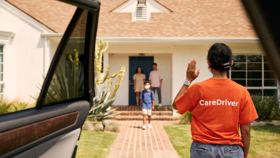 A HopSkipDrive caregiver waits by an open car door in front of a house.