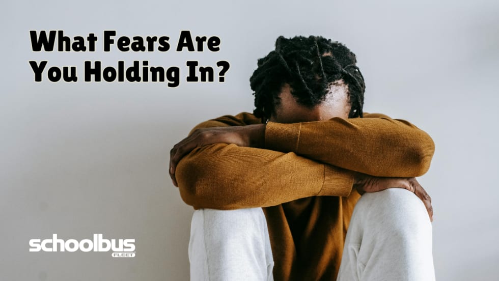 A person sits on the floor with their knees pulled in and arms crossed over them, head resting down on their arms, conveying stress or anxiety. Text on the image reads, “What Fears Are You Holding In?” with the School Bus Fleet logo in the corner.