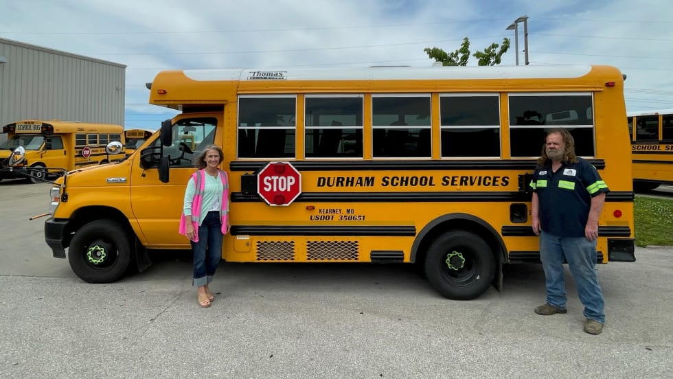 A Durham School Services bus with two people posing in front of it.