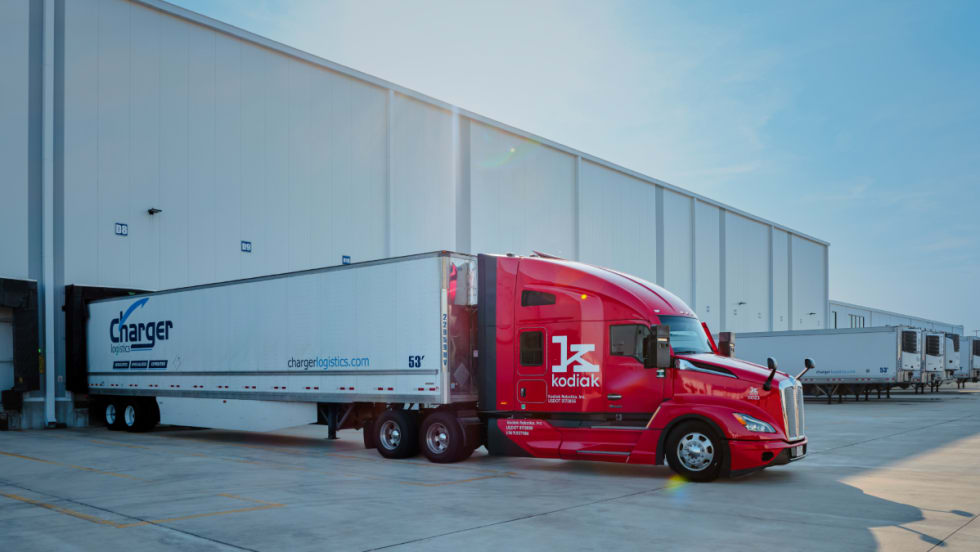 Red Kodiak-branded autonomous tractor with Charger-branded trailer backed up to warehouse