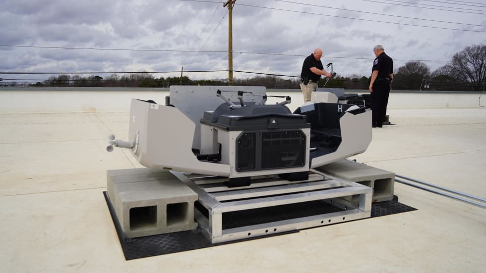 Two police officers stand near a rooftop drone launch system, preparing equipment for a first responder program providing aerial support to emergency calls.