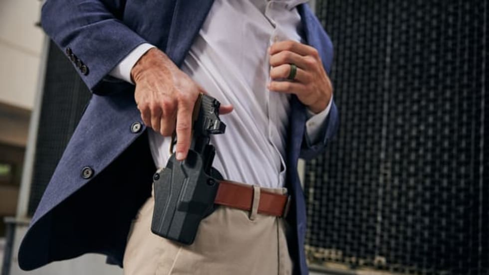 Man in a blue blazer lifting his shirt to reveal a concealed handgun in a waistband holster while standing outdoors.