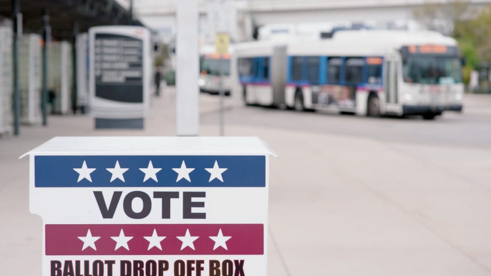 A ballot drop-off box marked “Vote” stands in the foreground at a transit station, with public transit buses visible in the background.