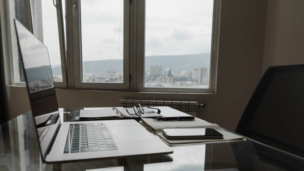Photo of a laptop, cellphone and eyeglasses on a desk next to an office window