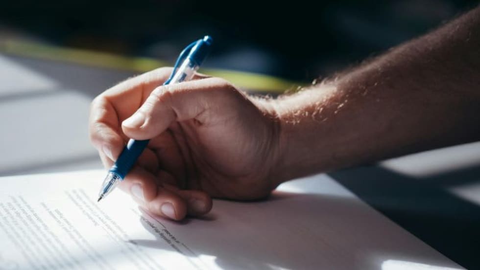 Photo of man's hand holding a pen above a piece of paper on a desk