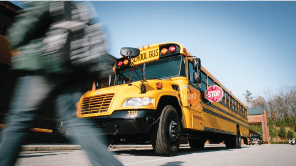 A yellow school bus with its red stop arm extended is stopped to load or unload students as a blurred child crosses in the foreground, illustrating the importance of motorists stopping for school buses and protecting students in the loading zone.