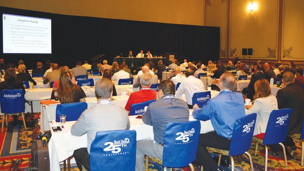Audience seated at tables attending an automotive industry conference session, with a panel of speakers presenting on stage and a presentation slide displayed on a large screen.