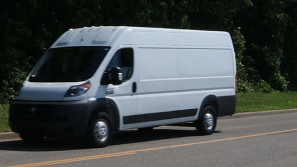 White cargo van driving on a paved road with trees and greenery in the background.