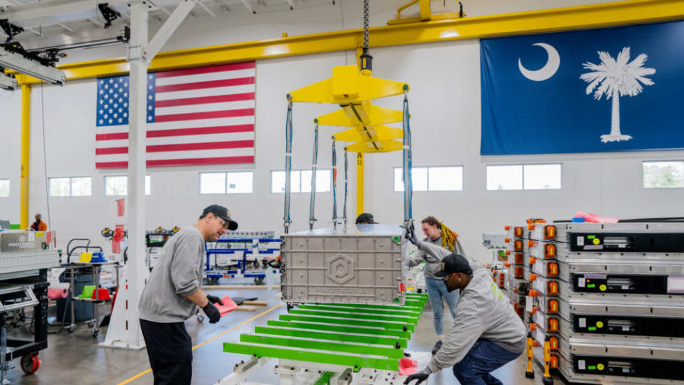 Workers assemble a large Proterra EV battery pack inside a manufacturing facility, using an overhead crane to position the battery module onto a chassis frame. American and South Carolina state flags hang above the production floor, with additional battery packs stacked nearby.