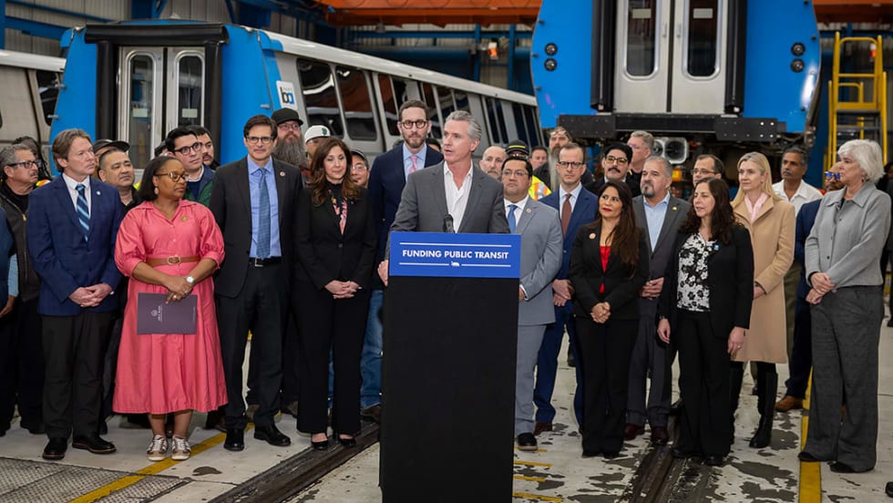 California Governor Gavin Newsom stands at a podium surrounded by a group of state and community leaders.