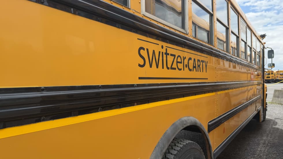 A close-up side view of a yellow Switzer-CARTY school bus parked in a bus yard, with the contractor's name displayed on the side panel and additional school buses visible in the background.