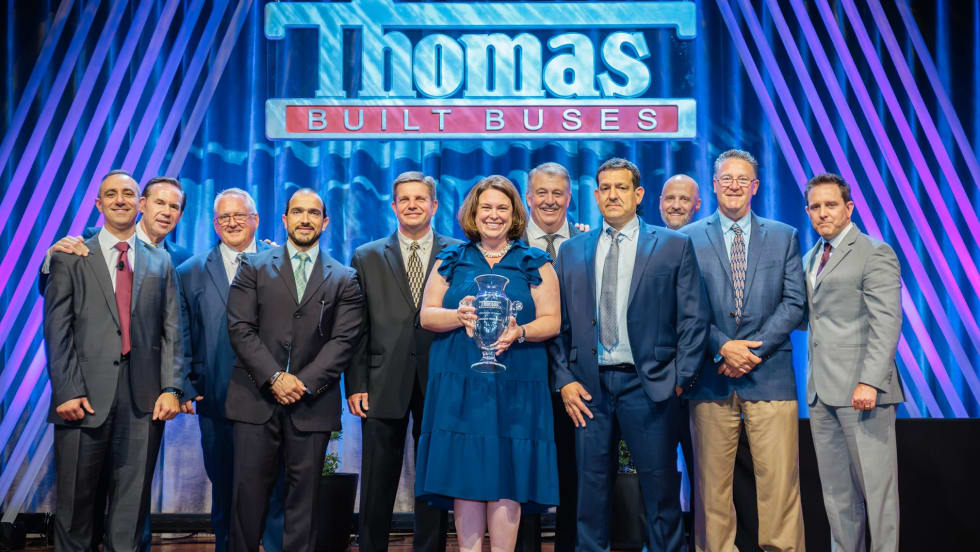 Group of Thomas Built Buses executives, employees, and award recipients pose onstage during an awards ceremony beneath a large Thomas Built Buses sign, with one woman holding a glass trophy at center stage.