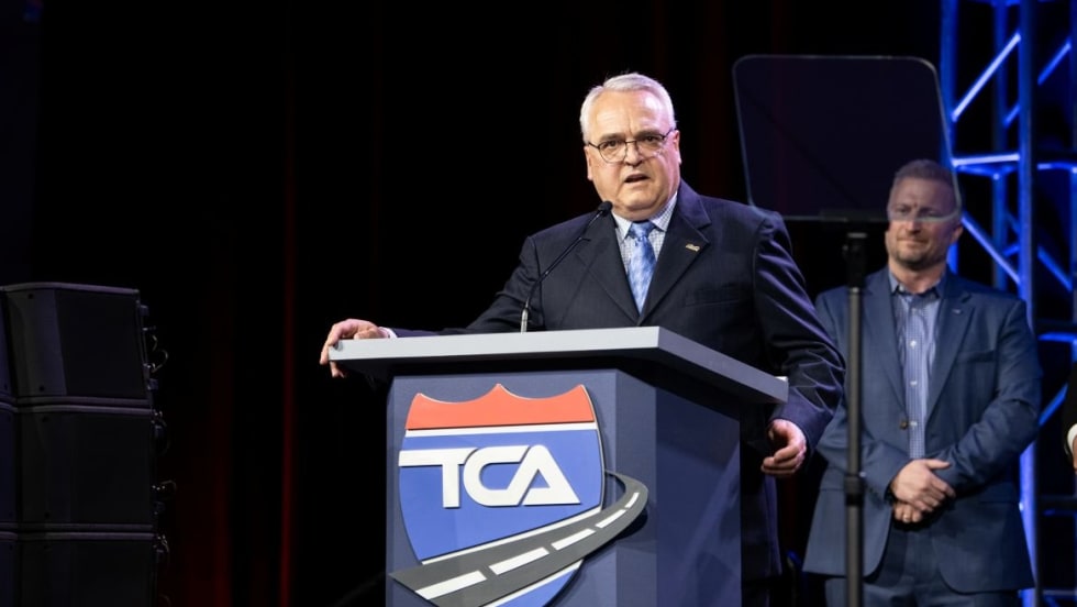 Older white man in suit standing at podium with TCA logo