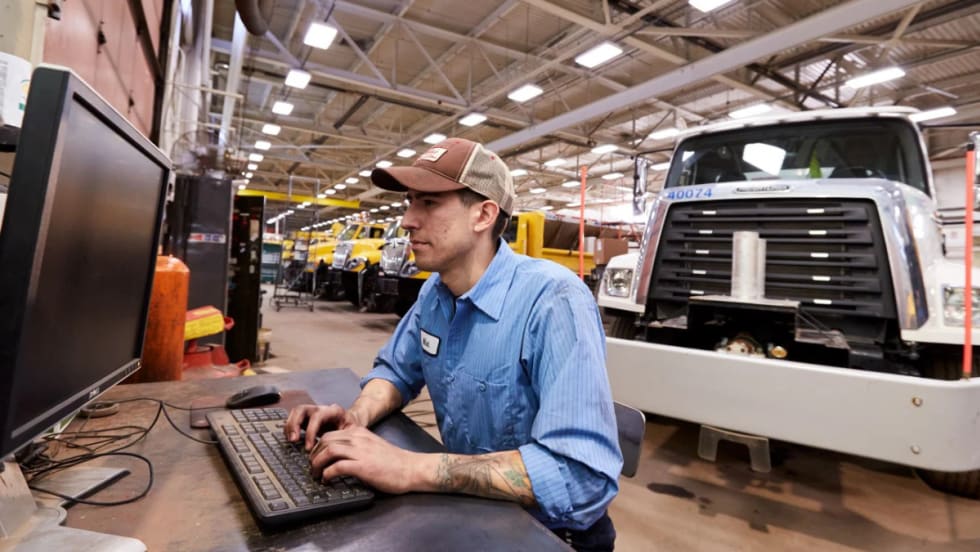 Technician at computer in maintenance shop with truck in background