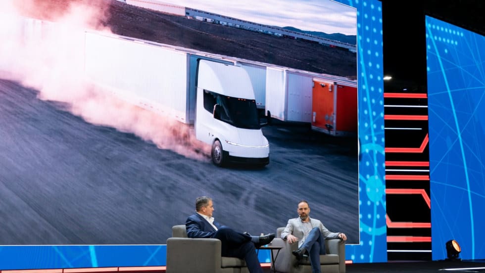 Two men in chairs on stage with video image of Tesla electric truck on big screen