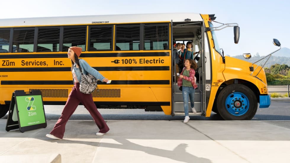 Students getting off a yellow electric school bus labeled “100% Electric” with Zūm Services branding
